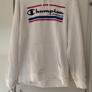 Vit Champion hoodie med logga - Klassisk Champion hoodie med snörning och stor magficka. Mjuk bomullskänsla utan att vara för tjock i materialet och långärmad modell. Liten Champion-logga på ärmen.