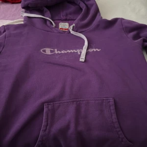 Lila Champion hoodie med snörning - Säljer en lila hoodie från Champion i storlek M. Tröjan har vit snörning vid huvan, stor magficka och broderad Champion-logga på bröstet. Perfekt för chill dagar och streetstyle. Materialet är mjuk bomull som känns skönt mot huden.