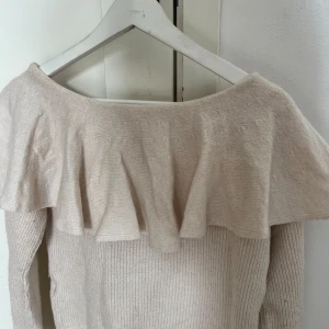 Beige stickad tröja med volang - Superfin beige stickad tröja med bred volang över axlarna. Tröjan har en offshoulder-design och är ribbstickad i materialet, vilket ger en snygg och trendig look. Perfekt för dig som gillar mjuka färger och detaljer som sticker ut.