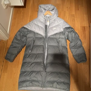 Nike jacka - Säljer en lång, grå dunjacka från Nike med Storm-FIT-teknologi. Jackan har tvåvägsdragkedja, huva och två sidofickor med dragkedja. Färgblockad i ljusgrått och mörkgrått, perfekt för kalla dagar. Klassisk pufferstil och riktigt skön passform.