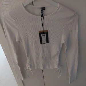 Ny Vit ribbad croppad topp från Pieces - Säljer en vit långärmad croppad topp från Pieces. Toppen är ribbad och har snygga knytband i sidorna för extra detalj. Perfekt för dig som gillar en enkel men trendig look. Rund halsringning och mjukt material gör den superbekväm. Från rök och djurfritt hem. 