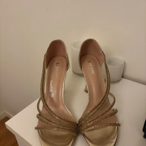 Guldiga klackskor  - Elegant och glittrig design  perfekta för bröllop, bal, fest eller andra speciella tillfällen. 	•	Storlek: 41 	•	Färg: Guld med glittrande remmar 	•	Öppen tå och bekväm klackhöjd ca 8 cm 	• aldrig använda endast provade.   Skorna sitter stabilt och ger en lyxig känsla till vilken outfit som helst.  💌 Skickas spårbart eller möts upp i Halmstad! 