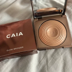 CAIA Glow Bronzer La Digue - Snygg bronzer från CAIA i nyansen La Digue. Kommer i en lyxig, bronsfärgad förpackning med spegel och har ett coolt cirkelmönster i pudret. Ger en varm, glowig finish och är perfekt för att skapa solkysst look. Pressat puderformat. Helt ny och aldrig använd endast öppnat förpackningen😊