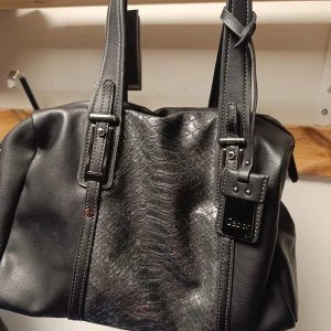 Svart handväska från Gabor i skinn - Snygg svart handväska från Gabor med coolt ormskinnsmönster på framsidan och detaljer i silver. Väskan har dubbla handtag, dragkedja bak och ett rymligt mönstrat innerfoder. Perfekt för dig som vill ha en stilren men ändå unik väska.