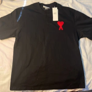 Svart t-shirt från Ami med rött hjärta - Svart t-shirt från Ami Alexandre Mattiussi med klassisk passform och korta ärmar. På bröstet finns ett broderat rött hjärta med bokstaven A, vilket ger en snygg och unik detalj. Materialet är mjuk bomull som känns skönt mot huden. Perfekt för dig som gillar stilrena plagg med en twist.