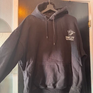 Svart hoodie Sweet Skateboards - Svart hoodie från Sweet Skateboards med stor vit print på ryggen av en ängel som spelar gitarr och musiknoter. Framtill finns mindre logga med samma motiv. Hoodien har huva med snörning och stor magficka. Perfekt för chill och streetwear.