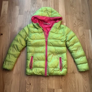 Grön pufferjacka med rosa detaljer - Snygg grön pufferjacka från Name It med rosa foder och dragkedjor. Jackan har huva, långa ärmar och två fickor framtill. Perfekt för kyliga dagar och ger en färgklick till din outfit. Passar dig som gillar att sticka ut med färg!har lite fläckar här och där men är annars i bra skick 