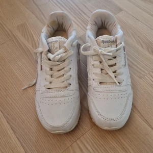 Vita Reebok Classic sneakers i skinn - Säljer ett par vita Reebok Classic sneakers i skinn med rund tå och platt sula. Skorna har snörning och diskreta perforeringar framtill för extra ventilation. Klassisk Reebok-logga på sidan och bak, samt Union Jack-detalj. Perfekta för en clean och sportig stil.