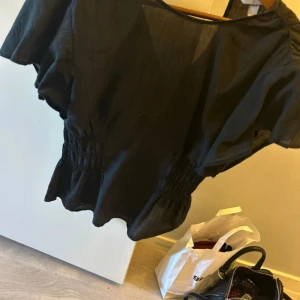 Svart blus med vida ärmar - Svart blus med breda, luftiga kortärmar och smockad midja som ger en snygg siluett. Tunt och lätt material, perfekt för en chill och stilren look. Passar till både jeans och kjol.