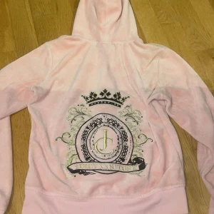 Rosa velour hoodie från Juicy Couture - Supermjuk rosa hoodie från Juicy Couture i velour med dragkedja, huva och känguruficka. Baksidan har stort glittrigt emblem med JC-logga och krona. Perfekt för en chill och trendig look.