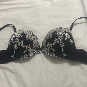 Svart spets-bh från Lindex - Snygg svart bh från Lindex i storlek 80A med vacker spets i vitt blommönster över kuporna. Dekorativ liten rosett framtill och tunna axelband. Perfekt för dig som gillar detaljer och vill ha något extra under kläderna. Helt ny och nästan aldrig använd!💗OBS, katt finns i hemmet!