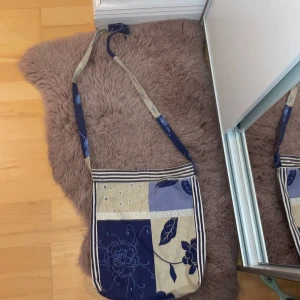 Handgjord axelväska från Sanna Design - Unik axelväska i patchwork-stil från Sanna Design. Väskan har blå, beige och vita toner med blommigt och randigt mönster. Lång axelrem och praktisk storlek för vardagens prylar. Tillverkad i bomullstyg med fina detaljer och knytstängning upptill.aldrig använd. Hör av dig om du har frågor eller funderingar 💕