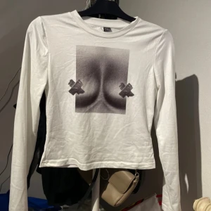 Vit långärmad topp med tryck från SHEIN - Vit långärmad topp från SHEIN i storlek S. Framsidan har ett edgy svartvitt tryck med bröstmotiv och tejpdetaljer. Modellen är croppad med rund hals och tillverkad i mjuk bomull. Perfekt för dig som gillar statement-plagg och vill sticka ut.