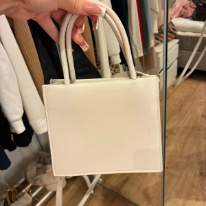 Beige fyrkantig handväska med axelrem - Säljer en stilren beige handväska i fyrkantig modell med både handtag och lång axelrem. Väskan är i syntetmaterial och har minimalistisk design utan mönster eller detaljer. Perfekt storlek för det viktigaste och passar till många outfits.