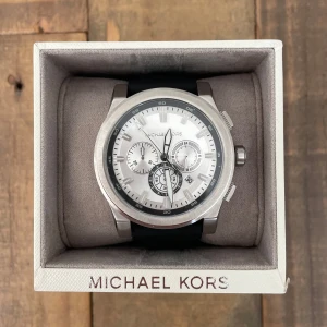 Michael Kors silverfärgad klocka - Stilren klocka från Michael Kors med silverfärgat rostfritt stål och vit urtavla. Klockan har flera små urtavlor och svart silikonarmband med klassiskt spänne. Perfekt accessoar för dig som gillar en clean och modern look. Det är nytt batteri och är helt ny i original förpackning men har varit oänvänd i tre år. Den är inte vattentät och är i modell MK8596 och nypriset är 3000kr.
