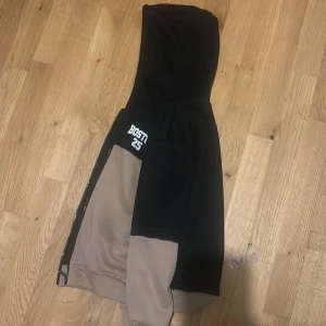 Svart och beige hoodie med BOSTI 25 - Säljer en snygg hoodie från BOSTI med trycket 'BOSTI 25' på bröstet. Tröjan har en cool färgblock-design i svart och beige, huva och dragkedja framtill. Perfekt för dig som gillar streetwear och vill ha en chill look. För en 8 åring