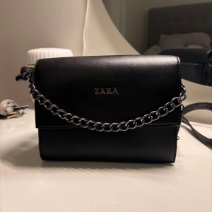 Svart axelväska med kedja - Snygg svart axelväska med stilren silverfärgad kedja som detalj. Väskan har ett magnetlås framtill, dragkedja på baksidan och framsidan och rymlig inuti för smidig organisering. Tillverkad i skinnimitation med en klassisk, boxig form och justerbar axelrem. Extra axelrem tillkommer. 