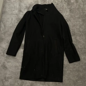 Svart kappa från Divided, stl S - Stilren svart kappa från Divided i storlek S. Klassisk rak modell med krage, dolda knappar och två fickor framtill. Perfekt för dig som gillar minimalistisk och clean look. Materialet är mjukt och känns skönt mot huden.