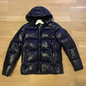 Svart pufferjacka från Save The Duck - Säljer en svart, glansig pufferjacka från Save The Duck i storlek M. Jackan har huva, två dragkedjefickor och är helt animal free. Köpt förra vintern från MIINTO för 3500 ungefär. Och den användes Max 10 gånger så har bara legat i garderoben sen dess. Skicket e 10/10. Mitt pris är 1999kr. Hör av er vid minsta!