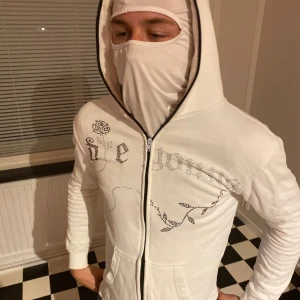 Vit hoodie med strassrosor och dragkedja - Vit hoodie med hel dragkedja och huva. Dekorerad med strassdetaljer i form av rosor och text framtill och på ryggen. Mjuk bomullskänsla, ribbade muddar och fickor framtill. Perfekt för dig som gillar unika detaljer och lite bling.