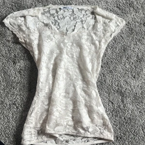 Vit spetsblus från Gina Tricot - Säljer en söt vit spetsblus från Gina Tricot i storlek XS. Blusen har korta ärmar, v-ringning och är helt täckt av blommig spets.
