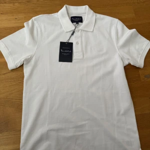 Vit pikétröja från Bondelid - Klassisk vit pikétröja från Bondelid med krage och knappar framtill. Tröjan har diskret broderad logga på bröstet och är tillverkad i mjuk bomull. Perfekt för en stilren och enkel look.