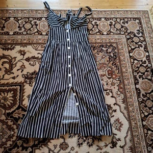 Svartvit randig klänning med knappar - Säljer en svartvit randig klänning med knappar framtill och smala axelband. Klänningen har en feminin passform med markerad midja och är i midi-längd. Perfekt för varma dagar och enkel att styla med sneakers eller sandaler.