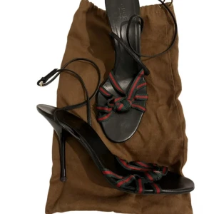 Svarta sandaler med klack från Gucci - Säljer ett par svarta sandaler med hög klack från Gucci. Skorna har remmar i grönt och rött tyg med knutdetalj framtill och smala svarta läderremmar runt vristen. Perfekta för dig som vill ha en lyxig och trendig look.