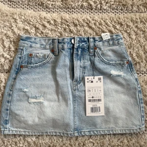 Ljusblå jeanskjol från Stradivarius - Säljer en ljusblå jeanskjol från Stradivarius, köpt på zalando. Kjolen är kort och har slitna detaljer framtill för en trendig look. Helt ny, säljer endast pga att den är för liten❤️ Jag har ca 40 cm rakt över midjan och den går precis inte att knäppa.