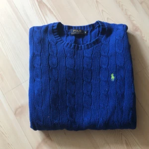 Blå kabelstickad tröja från Polo Ralph Lauren - Säljer en klassisk blå kabelstickad tröja från Polo Ralph Lauren. Tröjan har rund halsringning och den ikoniska broderade loggan i grönt på bröstet. Perfekt för dig som gillar tidlös stil och vill ha något som sticker ut lite extra. Perfekt för sena sommarkvällar eller annat!