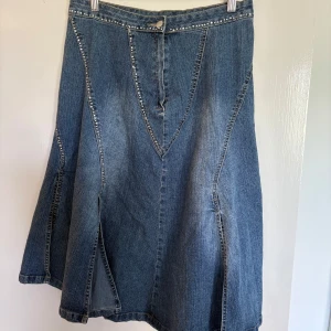 Jeanskjol med rhinestones - Säljer en snygg jeanskjol med rhinestones längs sömmarna och midjan. Kjolen har en A-linjeform och stängs med knapp och dragkedja framtill. 