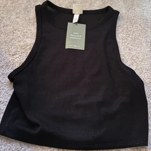 Svart croppad linnetopp från H&M - Säljer en svart glittrig croppad linnetopp från H&M, tillverkad av 60% återvunnen polyester. Toppen har en enkel och stilren design med rund halsringning och ärmlöst snitt. Perfekt att matcha med jeans eller kjol. Helt ny med prislapp