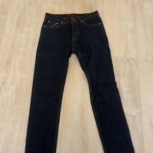 Mörkblå jeans från Tommy Hilfiger - Snygga mörkblå jeans från Tommy Hilfiger med orange sömmar. Modellen är Tapered Fit med avsmalnande ben och midwaist. Klassisk femficksdesign och diskreta detaljer i rött och vitt vid linningen.