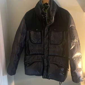 Svart pufferjacka från Hugo Boss - Väldigt bra skick. Säljer en svart pufferjacka från Hugo Boss med hög krage och stora fickor framtill. Jackan har quiltad design och stängs med dragkedja och knappar. Perfekt för kyliga dagar och har en stilren look med diskret logga på bröstet.