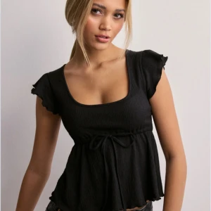 Svart ribbad topp med volangärm och knyt - Säljer en svart ribbad topp med volangärmar och söt knytdetalj i midjan från Nelly, säljer då den inte kommer till användning. Skriv för fler bilder eller andra frågor💕