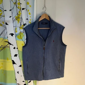 Blå väst från Polo Ralph Lauren - Snygg blå väst från Polo Ralph Lauren med dragkedja framtill och två fickor med dragkedja. Klassisk design med liten röd broderad logga på bröstet och hög krage. Perfekt inför sommarn eftersom den är lite tunnare. Nypris 2000kr. 