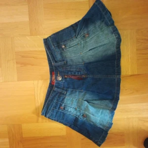 Jeanskjol från Voelle Denim - Säljer en blå jeanskjol från Voelle Denim med fickor fram och bak, dekorativa knappar och dragkedja. Kjolen har en kort modell och klassisk denimlook med ljusare tvättade partier. Perfekt för dig som gillar en avslappnad stil.