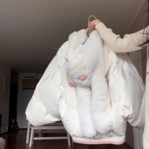 Vit pufferjacka med kaninhuva - Supergullig vit pufferjacka med stor fluffig kaninhuva och långa öron. Jackan har rosa detaljer och ribbad mudd nertill. Perfekt för dig som vill sticka ut och gillar söta detaljer.