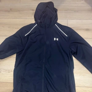 Svart vindjacka med huva från Under Armour - Har använt men ska tvätta innan skick, tvätt vänlig. Säljer en svart vindjacka från Under Armour med huva och dragkedja. Jackan har vita detaljer vid axlarna och logga på bröstet samt stor logotyp på ryggen. Perfekt för träning utomhus.