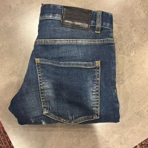 Blå jeans från Dsquared2 - Säljer ett par blå jeans från Dsquared2 med slitningar och lätt tvättad look. Jeansen har klassisk femficksdesign, gylf med dragkedja och knapp samt Dsquared2-lapp bak i midjan. Materialet är bomull med lite elastan för stretch. De har knappt blivit använda. De är slim modell. Hör av er om ni har frågor 