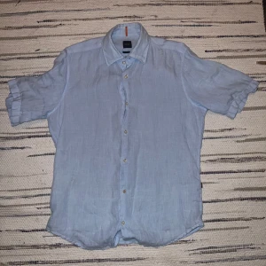 Ljusblå linneskjorta från BOSS - Säljer en ljusblå kortärmad skjorta från BOSS i linne. Skjortan har klassisk krage, knäppning framtill och en avslappnad regular fit. Perfekt för varma dagar och enkel att styla till olika outfits.