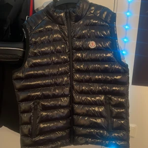 Svart Moncler väst  - Snygg svart dunväst från Moncler med glansig finish och diskret logotyp på bröstet. Västen har dragkedja framtill och två fickor. Perfekt för lager-på-lager och ger en sportig look.