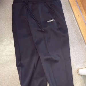 Svarta byxor från Les Deux med dragsko - Snygga svarta byxor från Les Deux med dragsko i midjan och diskret logga på ena benet. Har kvitto 