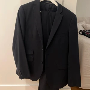 Svart kostym från Riley - Säljer en klassisk svart kostym från Riley. Kavajen har två knappar, smala slag och fickor med lock. Tillhörande byxor i samma svarta tyg. Storlek M