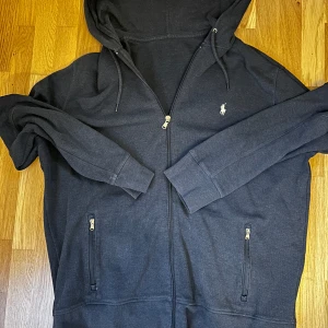 Ralph Lauren Hoodie - Säljer en mörkblå hoodie från Ralph Lauren med dragkedja och huva. Tröjan har två fickor framtill med dragkedja och den klassiska loggan broderad på bröstet. Perfekt för en avslappnad stil.