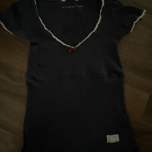 Marinblå ribbad tröja med spetsdetaljer - Söt marinblå ribbad tröja med vit spets längs v-ringningen och ärmsluten. Liten söt rosett framtill vid halsen. Kortärmad och figurnära modell, perfekt för en avslappnad look. Den har en liten fläck som säkert går bort i tvätten 🌸😊