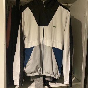 Vit och svart vindjacka från Lacoste - Säljer en snygg vindjacka från Lacoste i vitt, svart och blått. Jackan har en dragkedja framtill och en broderad krokodil-logga på bröstet. Perfekt för vår och höst med sin lätta och bekväma passform. Den har långa ärmar och en huva för extra skydd mot vinden. Passar både till vardags och träning.