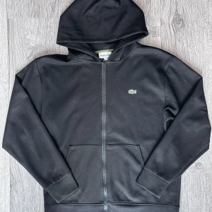 Lacoste Zip Hoodie Svart - Hej, säljer min svarta huvtröja med dragkedja från Lacoste. Tröjan är i mycket fint skick. Passar mycket bra. Etiketten som sitter på blixtlåslöparen har lossnat, se bild 3. (Detta påverkar inte blixtlåsmekanismen på något sätt). Hör av er vid frågor! 😄