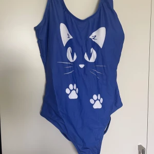Blå baddräkt med kattmotiv - Säljer en blå baddräkt med vitt kattansikte och tassar framtill. Baddräkten har djup rygg och breda axelband. Perfekt för dig som gillar djurmotiv och vill sticka ut på stranden.Helt ny