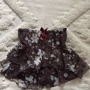 Brun blommig volangkjol / topp med rosett - Supersöt brun kjol med vita och ljusblå blommor, volanger och en vinröd rosett framtill. Passar st S-M. Resår i midjan för extra komfort. Vintage och köpt i Frankrike! 